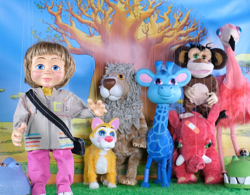 Petit Jean et les marionnettes du spectacle — girafe bleue, lion, singe, flamant rose — posant devant le décor